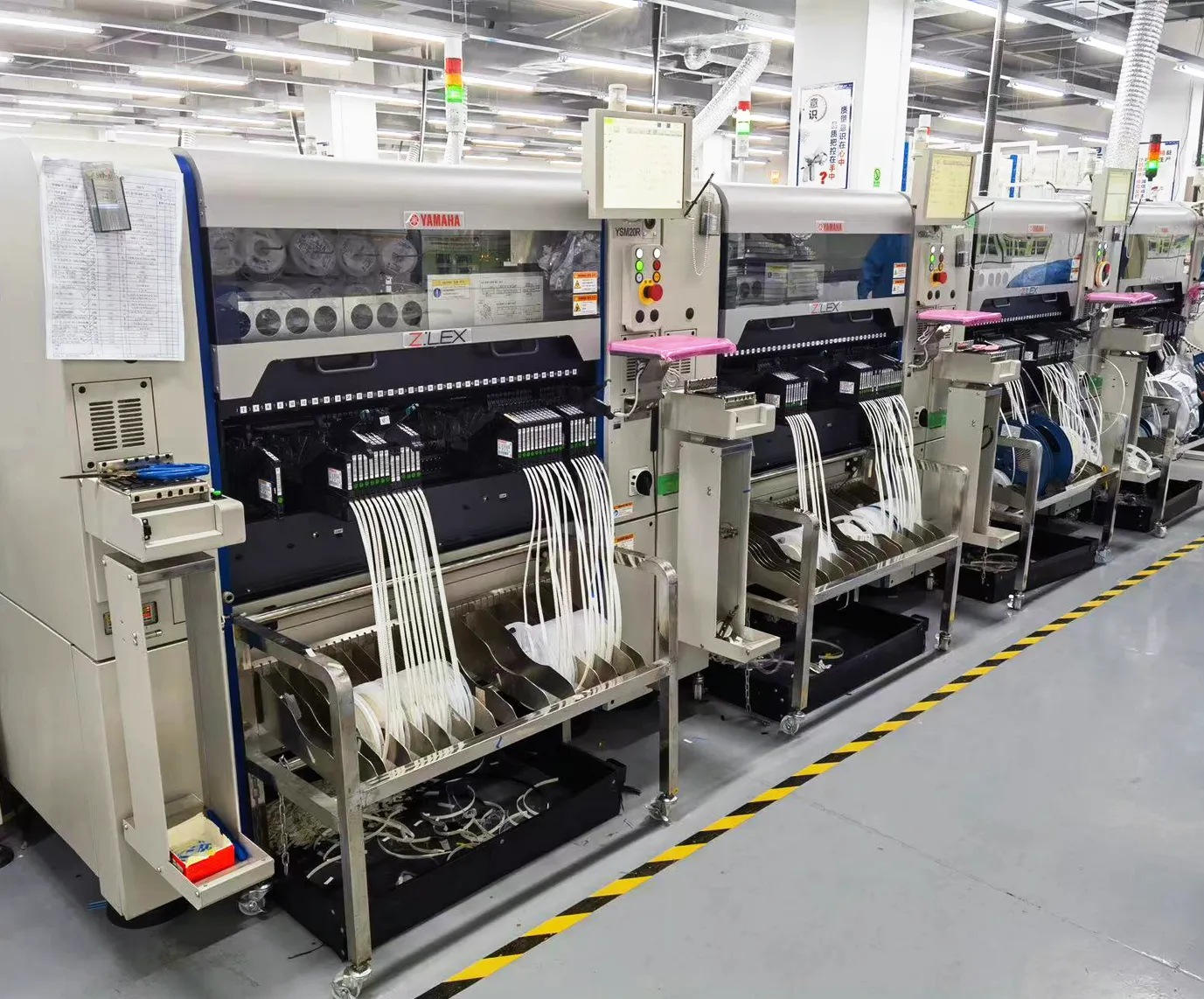 A row of Yamaha YSM20R mounters with feeder carts in a factory, white tape feeders loaded across the machines